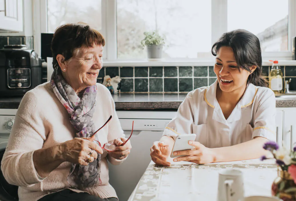 An elderly woman with short hair and glasses engages with a younger woman at a kitchen table. The younger woman, wearing a sweater, holds a smartphone, demonstrating it to the elder. They are surrounded by a homey kitchen interior, suggesting an informal health app consultation.